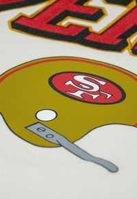 Goldener Football-Helm mit einem roten Streifen und Logo sowie weißen Akzenten am Kinnriemen, vor einem weißen Hintergrund mit fettgedrucktem roten Schriftzug.