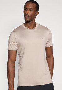 Μπεζ κοντομάνικο μπλουζάκι από απαλό ύφασμα, με στρογγυλή λαιμόκοψη και μικρό λογότυπο στο πάνω μέρος του στήθους.