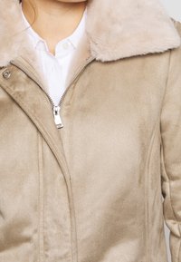 Chaqueta de ante beige con cuello de piel sintética, que presenta una cremallera frontal y una textura lisa. El diseño es ajustado con detalles de costuras.