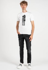 Camiseta blanca con una impresión negra que dice "LES HOMMES 1 EST. 2005," combinada con unos jeans negros de corte slim y zapatillas blancas con detalles en negro.
