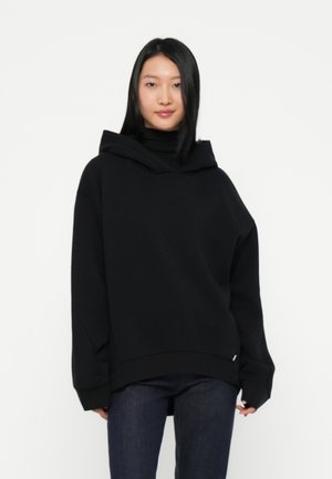 Felpa nera oversize con cappuccio profondo e maniche lunghe, realizzata in tessuto morbido, abbinata a jeans in denim scuro.