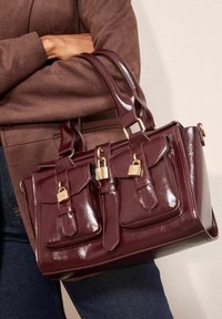 Burgundijas mākslīgās ādas rokassoma ar divām priekšējām kabatām, zelta aparatūras aksesuāriem un divām rokturiem. Struktūrēta virsma ar spīdīgu apdari.