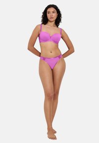 Vrouw die blootsvoets met krullend haar een bijpassende paarse beha en slipje modelleert tegen een plain witte achtergrond.