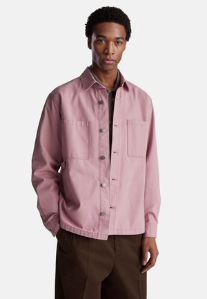 Roze katoenen jacket met twee grote voorzakken, sluiting met drukknoop en een ontspannen pasvorm. Gepaard met een bruine shirt eronder.