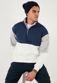 Tumesinise ja valge värviplokiga pullover, millel on veerandtõmble, hallid varrukad ja soonikäär. Kanga tekstuur näib pehme ja tekstureeritud.