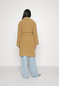 Cappotto color cammello con finitura testurizzata, maniche lunghe e cintura in vita, abbinato a jeans a gamba larga azzurro chiaro e calzature marroni.