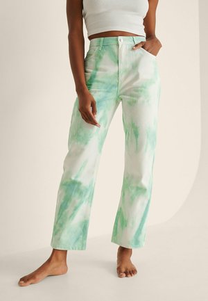 Kvinde iført højtaljede, løstsiddende jeans med grønne og hvide tie-dye mønstre og en hvid crop top, stående barfodet.