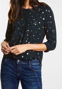 Svart blus med gyllene prickar, långärmad med uppkavlade ärmar, knappslå, lätt tyg, bärs tillsammans med blå jeans.