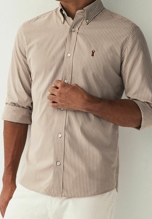 Homme portant une chemise à rayures verticales beiges et blanches à boutons, manches retroussées, et un pantalon blanc, main reposant près des boutons de poitrine.