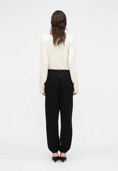 Γυναίκα με μακριά κυματιστά μαλλιά, φοράει λευκό μακρυμάνικο τοπ, μαύρο φαρδύ παντελόνι με ελαστικά τελειώματα και μαύρα παπούτσια με τακούνι, γυρισμένη με την πλάτη.