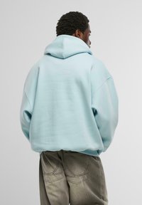 Sudadera oversize de color azul claro con capucha de doble capa, textura suave y hombros caídos, combinada con pantalones grises con costuras acentuadas.