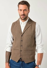Braun gemusterte Weste im Fischgrätenmuster mit fünf Knöpfen, Reverskragen und einer Brusttasche, über einem weißen Hemd. Kombiniert mit blauen Jeans.