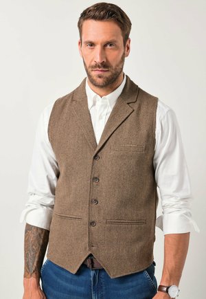 Braun gemusterte Weste im Fischgrätenmuster mit fünf Knöpfen, Reverskragen und einer Brusttasche, über einem weißen Hemd. Kombiniert mit blauen Jeans.