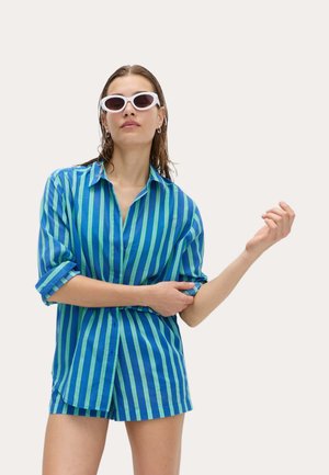 Femme portant une chemise rayée bleue et verte à boutons et un short assorti, ajustant la manche de la chemise, avec des lunettes de soleil blanches et les cheveux mouillés.