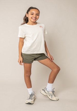 Jong meisje met paardenstaart, gekleed in een wit T-shirt, olijfgroene korte broek, witte sokken en grijze hardloopschoenen, staand tegen een beige achtergrond.