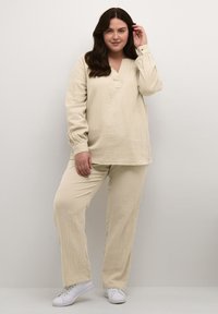 Helles beige Loungewear-Set featuring ein langärmliges Oberteil mit V-Ausschnitt und lockeren Hosen. Texturiertes Material und lässige Passform. Weiße Sneakers getragen.