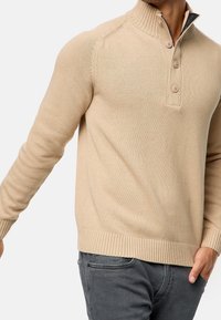 Beige gebreide trui met een hoge kraag, een sluiting met drie knopen en geribbelde mouwen en zoom. Gemaakt van zachte, getextureerde stof.