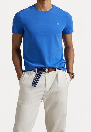 Blaues Baumwoll-T-Shirt mit Rundhalsausschnitt und kurzen Ärmeln, mit einem kleinen weißen Logo auf der Brust; kombiniert mit hellbeigen Hosen und einem gestreiften Gürtel.