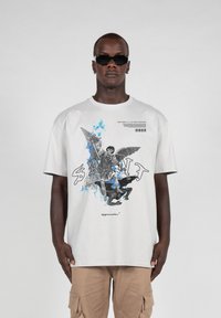 Hvid oversized t-shirt med et grafisk print af en engel og en figur i grå og blå nuancer, med tekstdetaljer og en afslappet pasform.