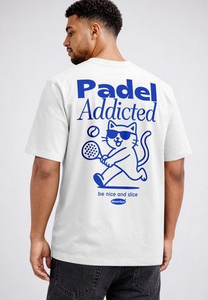 Man wearing white t-shirt with blue cat playing padel graphic and text "Padel Addicted" and "be nice and slice" on the back.