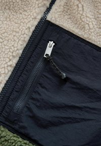 Fleecejakke med en sandfarvet fleeceoverdel, mørkeblå nylonbund og en lynlåslomme med sølvhardware samt en sort snor.