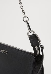 Sac en cuir noir avec une finition texturée, doté d'une fermeture éclair en métal argenté et d'une bandoulière en chaîne. Logo embossé sur le devant.