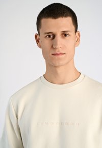 Krämfärgad sweatshirt med texturerad yta, prydd med en präglad "LINDBERGH"-logotyp på bröstet och rund halsringning.