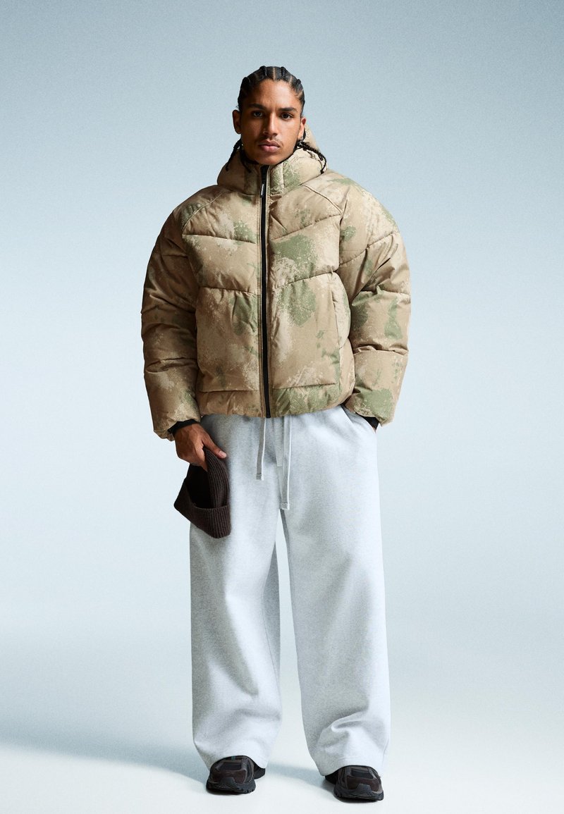 Khaki välsydd pufferjacka med en texturerad, mönstrad design. Kombinerad med grå sweatpants och håller en brun mössa. Mörka sneakers fulländar looken.