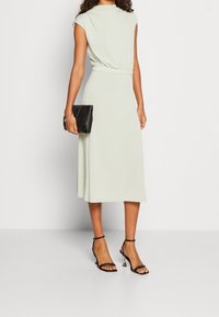 Ljusgrön midiklänning med korta ärmar och samlad midja, kombinerad med en svart kuvertväska och svarta högklackade sandaler. Slät tygtextur.