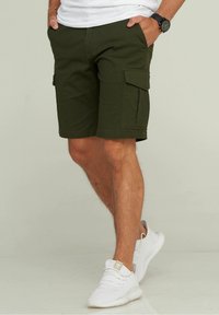 Shorts cargo verts olive avec poches latérales, fabriqués en tissu résistant, présentant une coupe droite et un ourlet, associés à des baskets blanches.