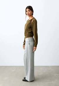 Mujer con camiseta de manga larga verde oliva con abertura en el hombro y vaqueros anchos de pierna gris claro, de pie sobre un suelo gris frente a una pared blanca.