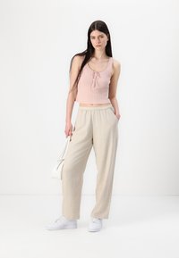 Top scurt cu dungi, de culoare roz deschis, cu detalii legate; pantaloni largi din in bej; teniși albi; ținând o geantă albă; fundal minimalist.