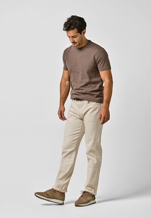 Hombre con camisa marrón de manga corta, pantalones crema y zapatillas marrones, mirando hacia abajo su pie izquierdo levantado sobre un fondo gris liso.