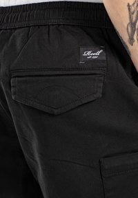 Pantalones negros con una tela texturizada, que presentan un bolsillo trasero con una solapa curva y una etiqueta de la marca que dice "Reell est. 1991."