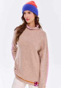Beige übergroßer Pullover mit rosa und orangenen Seitestreifen sowie einem rosa Kragen. Kombiniert mit einer bunten gestreiften Mütze. Weiche Textur, lockere Passform.