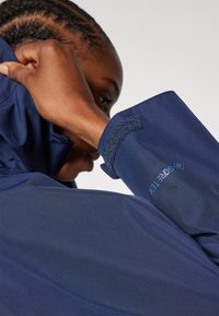 Navy waterproof jacket featuring a high collar, Velcro cuff adjustments, and a logo detail on the sleeve. Smooth, textured fabric.