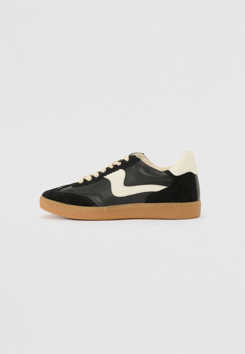 Sapatilha em camurça e couro pretos com um swoosh lateral em creme e um calcanhar em creme claro. Apresenta atacadores bege e uma sola de borracha tipo gum.