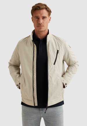 Mann trägt beige Zip-Up-Jacke über schwarzem Hemd und hellblauen Jeans, steht mit Händen in den Jackentaschen vor einfarbigem Hintergrund.
