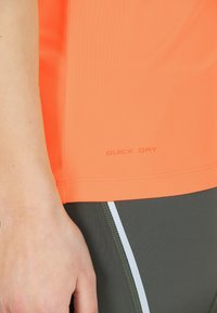 Lähikuva käsivarresta, joka on oranssin nopeasti kuivuvan paidan ja tummanharmaiden urheiluleggingien vieressä, joissa on valkoinen raita.