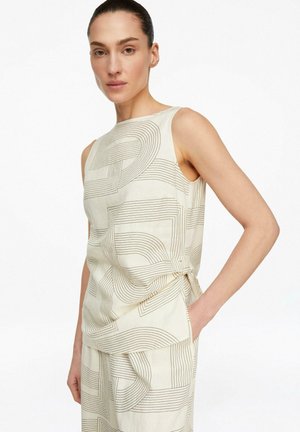 Kobieta w beżowej bluzce bez rękawów i spodniach z abstrakcyjnym wzorem liniowym, ręka w kieszeni, neutralny wyraz twarzy, jednolite białe tło.