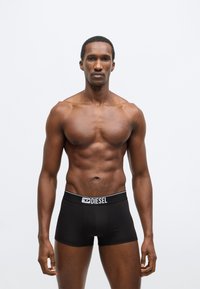 Fekete boxeralsó fehér logós derékpánttal, sima anyagból. A modell egyenes testtartásban áll, egyszínű háttér előtt.