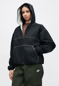 Μαύρο μπουφάν με φερμουάρ, με εσωτερική φόδρα κουκούλας σε γκρι, λευκή ραφή αντίθεσης και ελαστικά μανίκια· φορεμένο πάνω από καφέ μπλούζα και χακί παντελόνι.