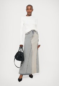 Stående kvinne med hvit langermet topp, to-farget grå denim maxiskjørt, svarte spisse sko, og holder en svart håndveske.