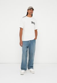 T-shirt bianco a maniche corte con stampa grafica nera e jeans denim blu relax. Accessoriato con un cappellino grigio chiaro e sneakers bianche.