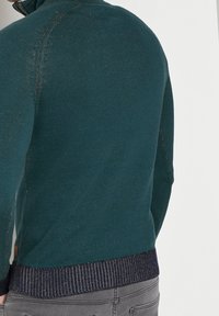 Maglione lavorato a maglia turchese con collo alto, orlo a coste blu navy, trama sottile e cuciture a contrasto, che evidenziano un design aderente.