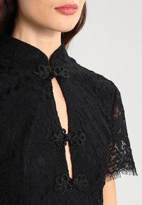 Black lace dress with short sleeves, mandarin collar, and decorative knot closures at the neckline, featuring a floral pattern throughout.