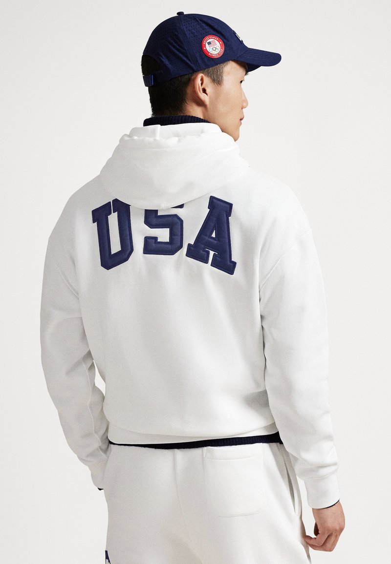 White hoodie with navy "USA" lettering on the back. Material appears soft, featuring a hood and ribbed cuffs. Navy cap with logo detail.
