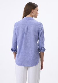 Camicia button-up in chambray blu con maniche lunghe, polsini arrotolati, dettaglio del giogo posteriore e orlo curvo. Tessuto leggero con una finitura testurizzata.