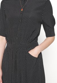 Czarna sukienka w białe kropki, guziki z przodu, rękawy do łokcia, elastyczna talia, kieszeń boczna z ręką w środku oraz warstwowy naszyjnik.