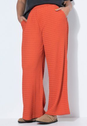 Persoon draagt textuurrijke, wijde, feloranje broek met handen in de zakken en beige instapschoenen tegen een witte achtergrond.